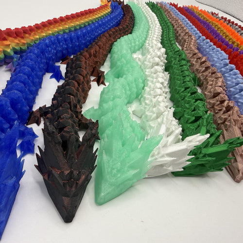A collection of 3D-printed articulated dragon figurines arranged in parallel rows, showcasing different color options including rainbow, blue, brown, mint green, white, forest green, and others.