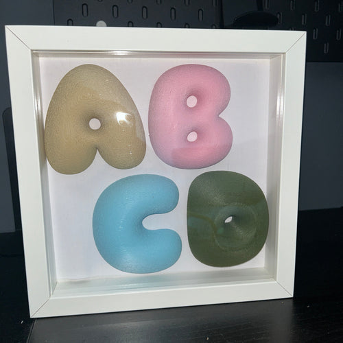 A white square shadow box frame displaying four stylized lowercase letters in beige, pink, light blue, and olive green, arranged to spell 'abco' on a white background.