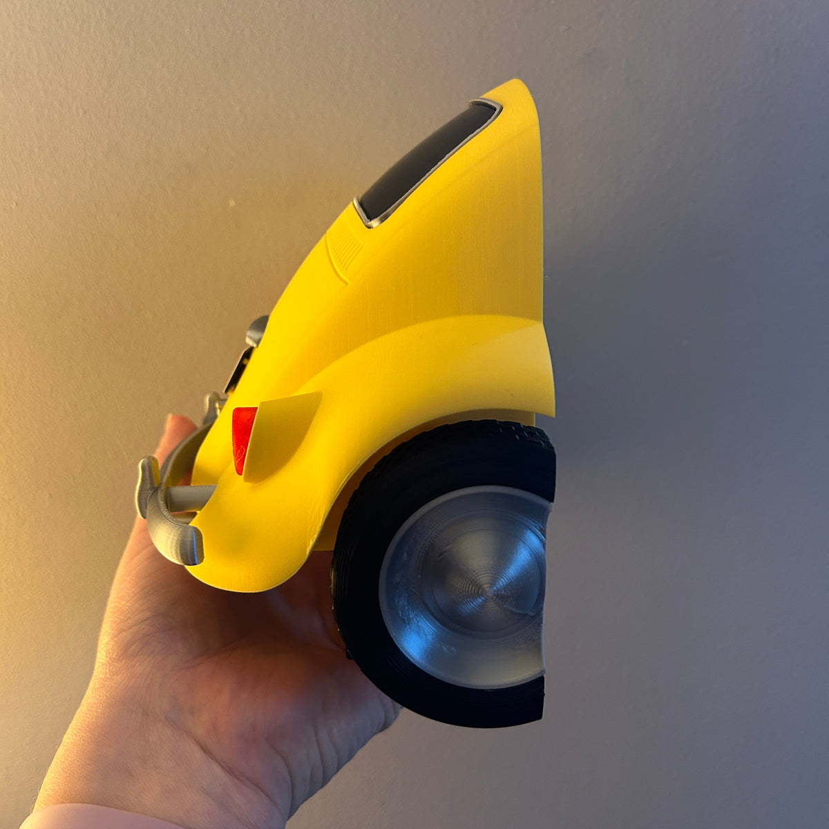 A hand holding a bright yellow car-shaped object with a black wheel and metallic silver center, photographed against a plain beige wall.
