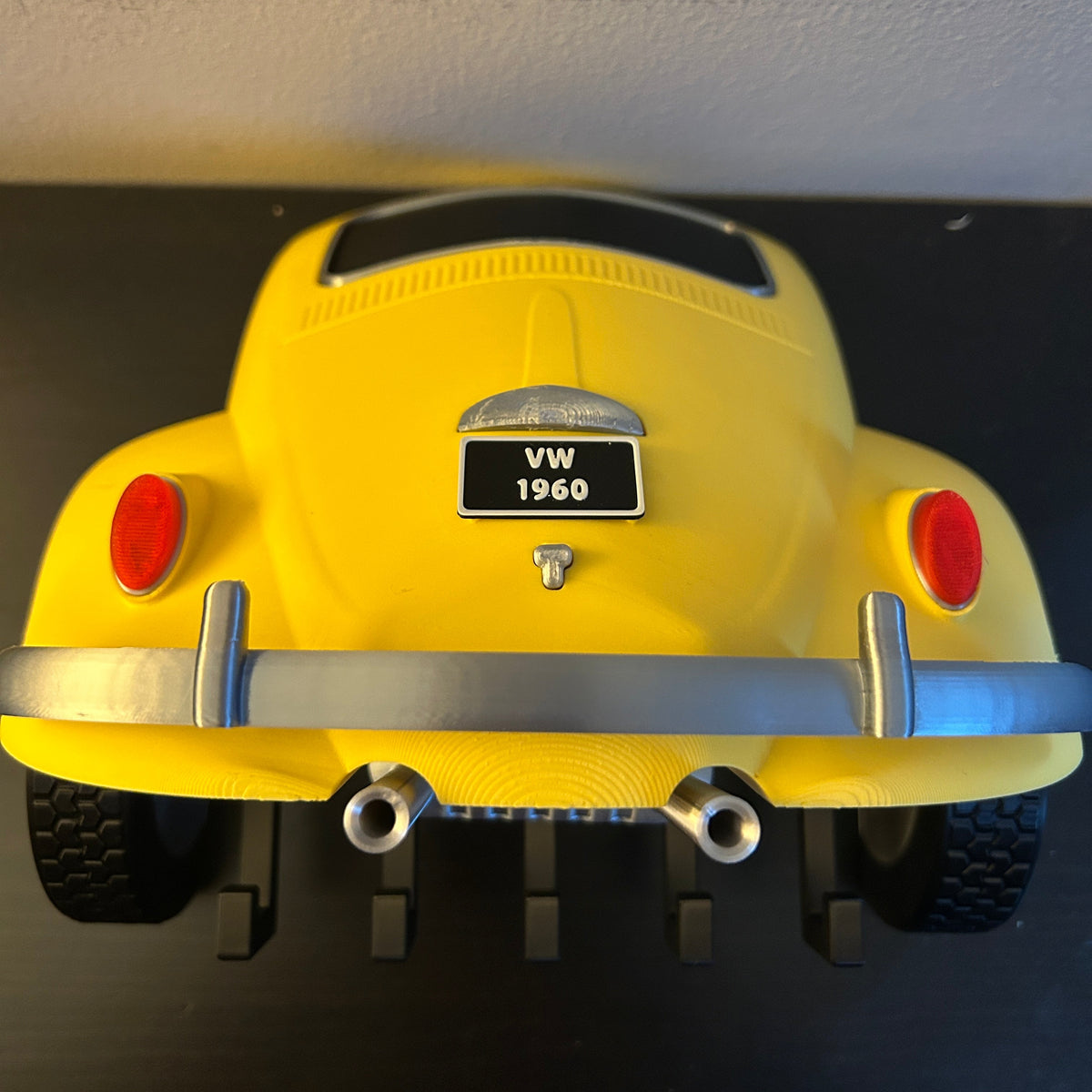 A yellow Beetle toy car shown from the rear, featuring five hooks for keys, a black license plate and a chrome bumper, displayed on a black surface against a light-coloured wall.