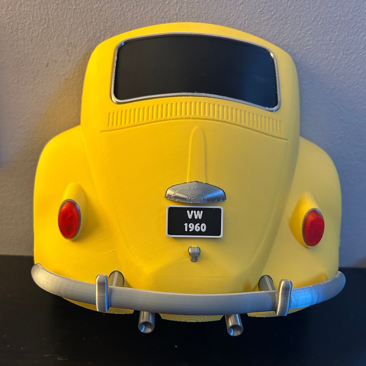A yellow vintage Beetle model for keys shown from the rear, featuring a black license plate reading and silver bumper details, set against a plain wall and dark surface.