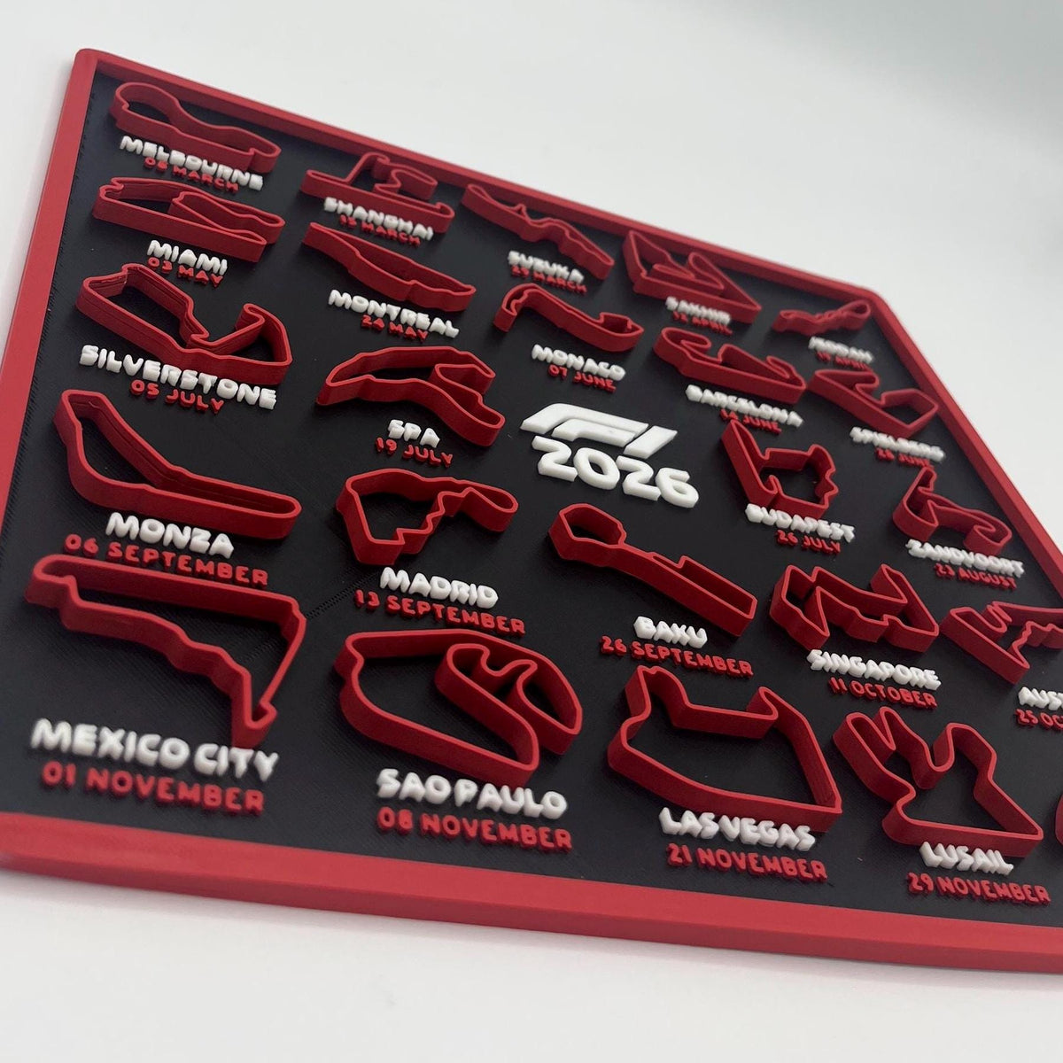 A 2026 F1 World Championship wall plaque featuring raised red and white race track silhouettes with city names and dates around the border, plus a central 2026 logo on a black background.