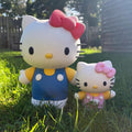 Two Hello Kitty garden statues in outdoor grass, one larger figure wearing blue overalls with a pink bow, and one smaller figure wearing pink polka dots with a pink bow.