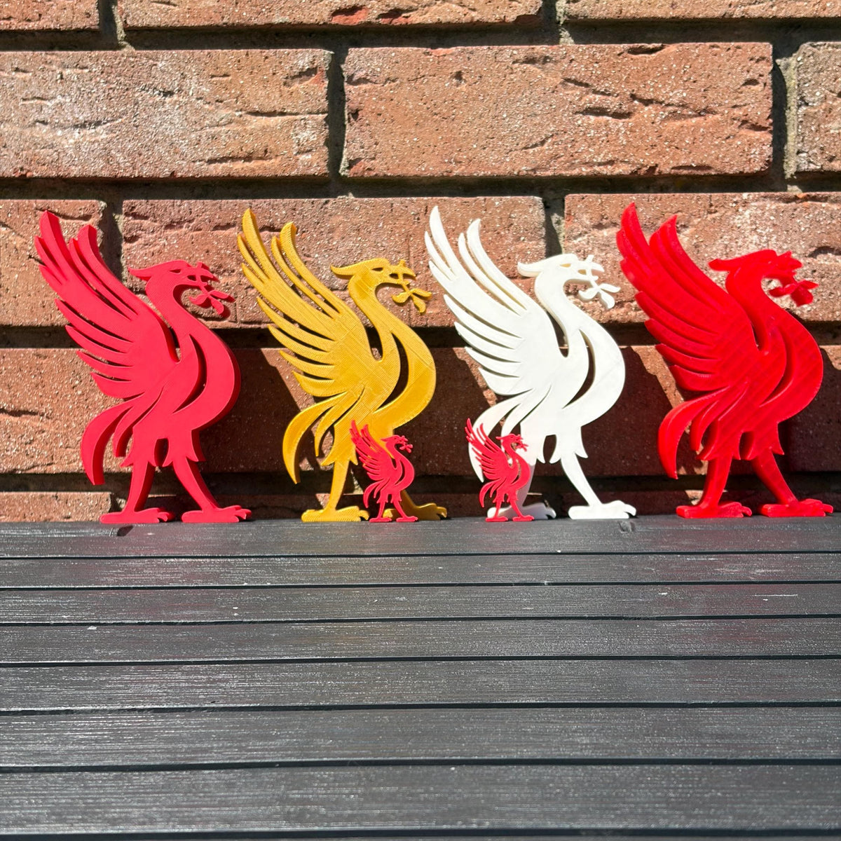 Six Liverpool bird 3D printed decorations in red, yellow, and white, arranged in a row on a dark wooden surface against a brick wall background.