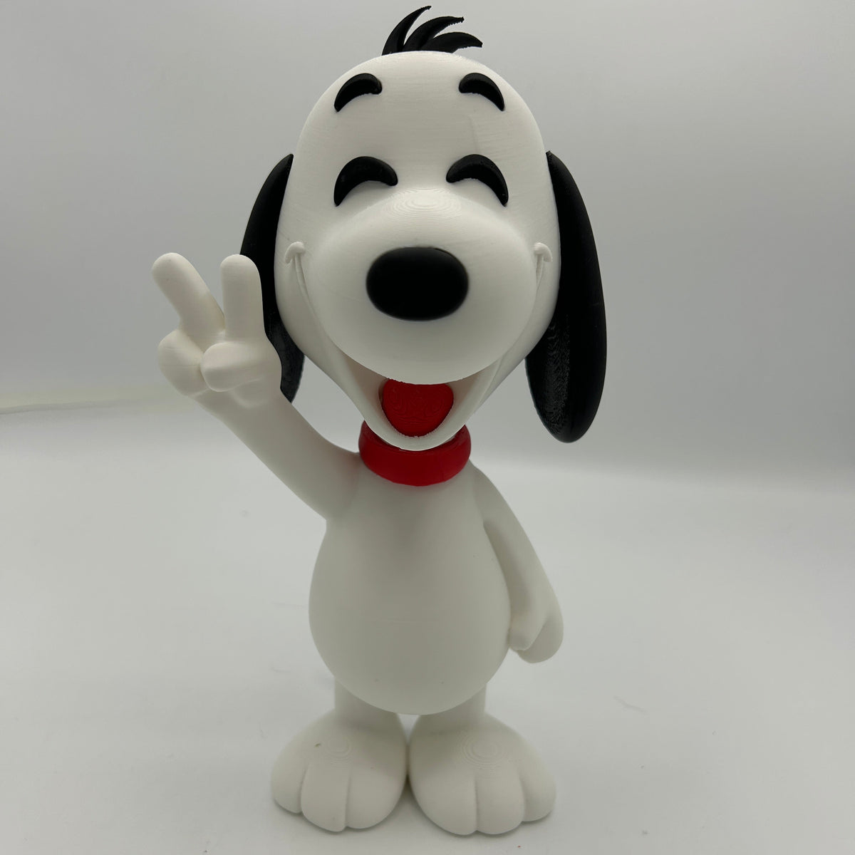 A white and black Snoopy figurine wearing a red collar, standing on a white surface against a light grey background and making a peace sign with its right paw.