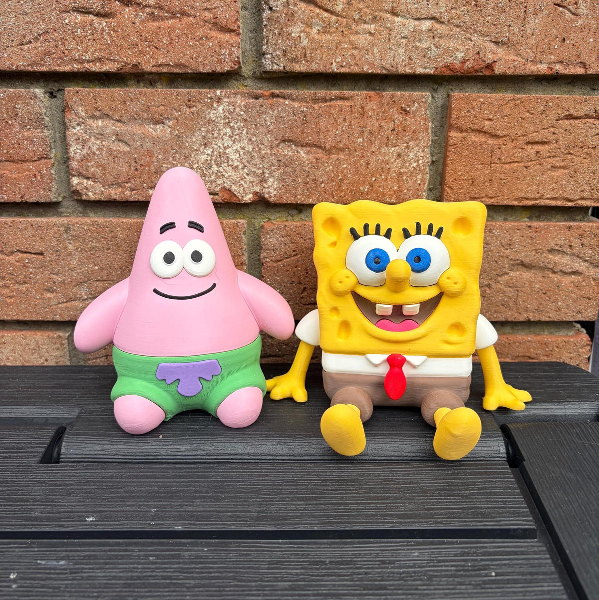 Two SpongeBob-themed 3D printed figurines, Patrick Star in green shorts and SpongeBob SquarePants in his white shirt with a red tie, sitting side-by-side on a dark wooden surface in front of a brick wall.