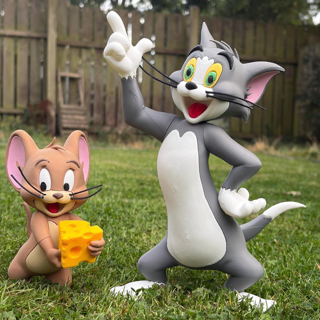 Tom and Jerry figurines in an outdoor backyard setting, with Tom the cat standing on grass and holding his paw out to point upward, and Jerry the mouse standing beside him holding a yellow cheese block.