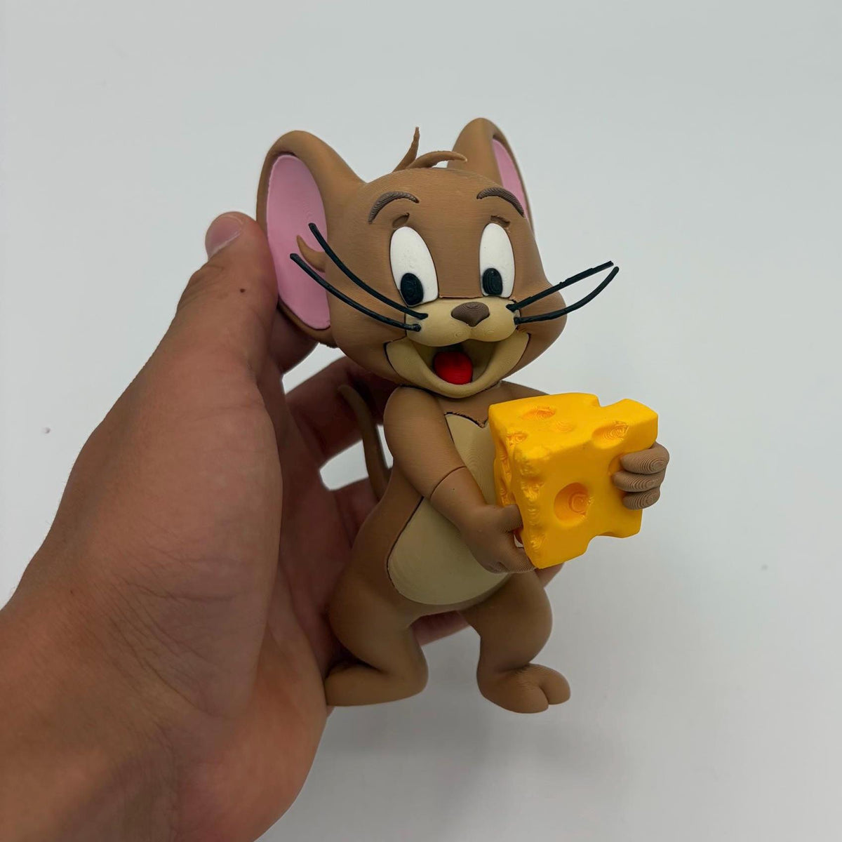 A hand holding a small Tom and Jerry figurine of Jerry holding a yellow cheese block, against a plain white background.