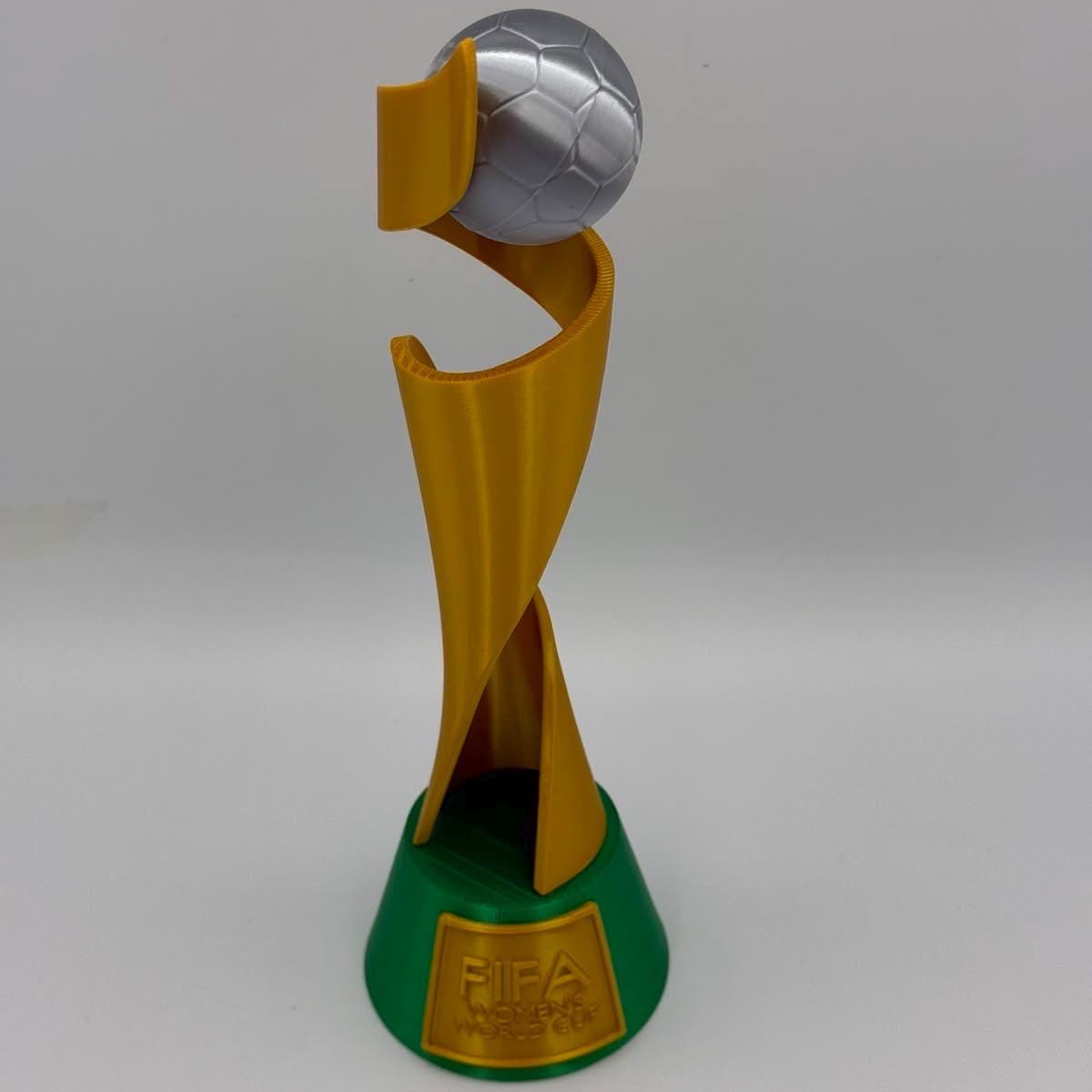 A replica of the FIFA Women's World Cup trophy, featuring a gold spiral structure holding a silver soccer ball, mounted on a green base with a gold FIFA World Cup plaque.