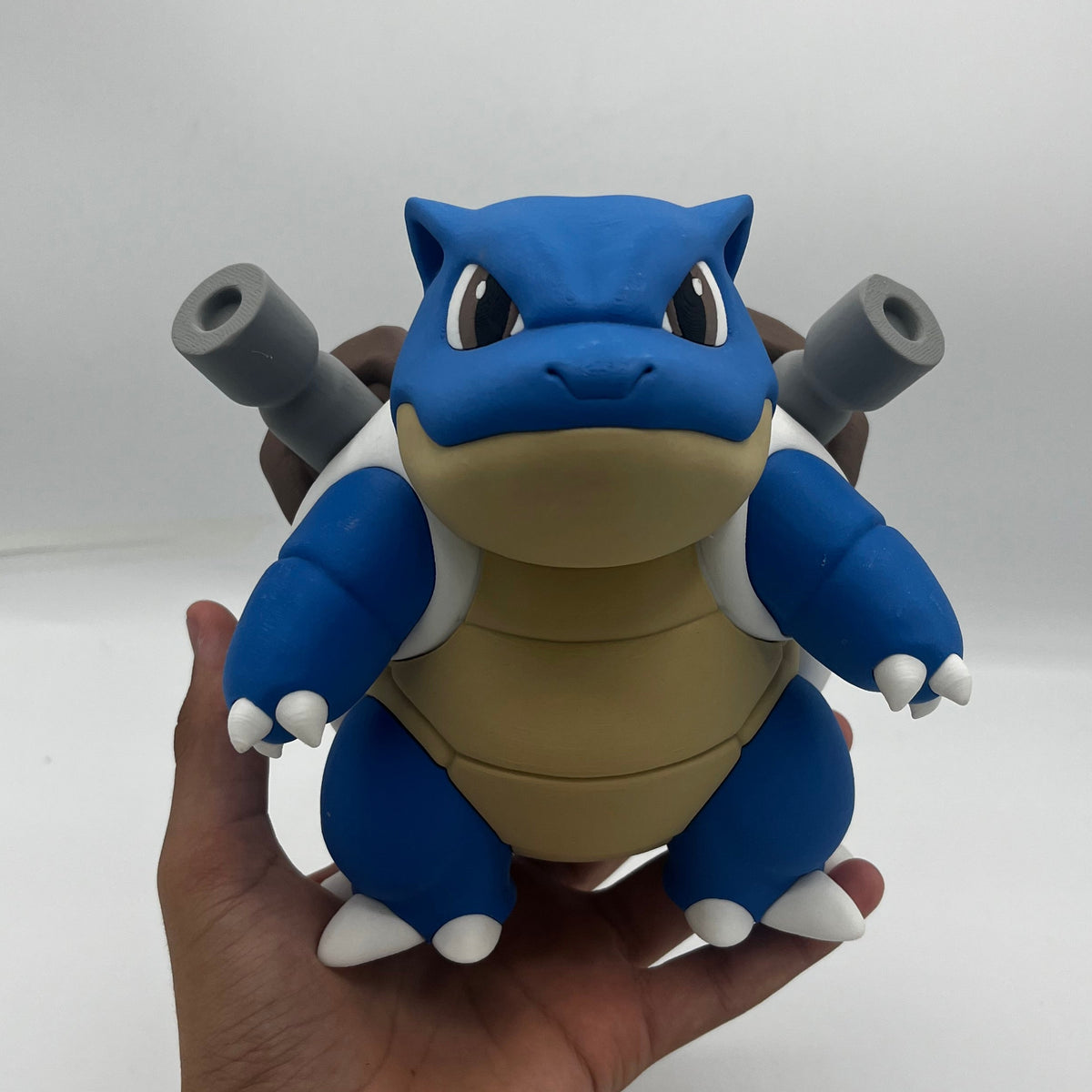 A blue and beige cartoon toy figure resembling a turtle-like character with gray cannon-like details on its shell, being held in a person's hand against a plain light background.