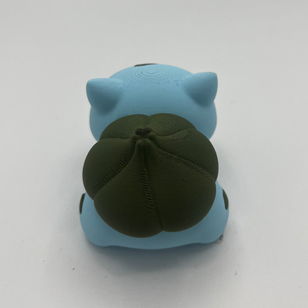 A light blue toy figurine shown from behind, featuring a dark olive green tail, photographed on a plain white background.