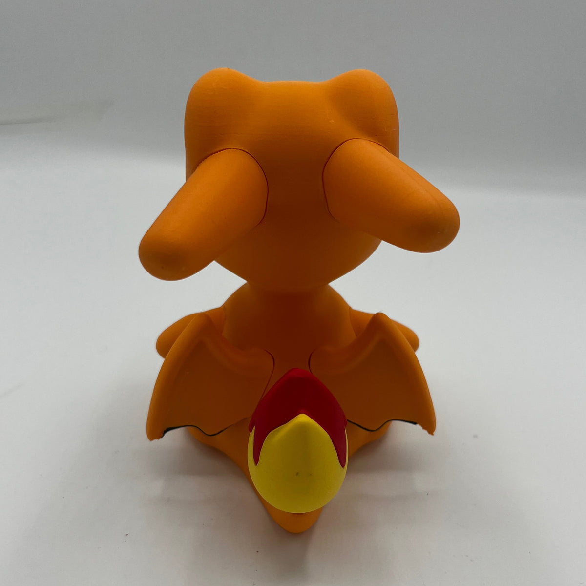 An orange toy figurine resembling a small dragon or bird character, photographed from the back on a plain light gray to white studio background.
