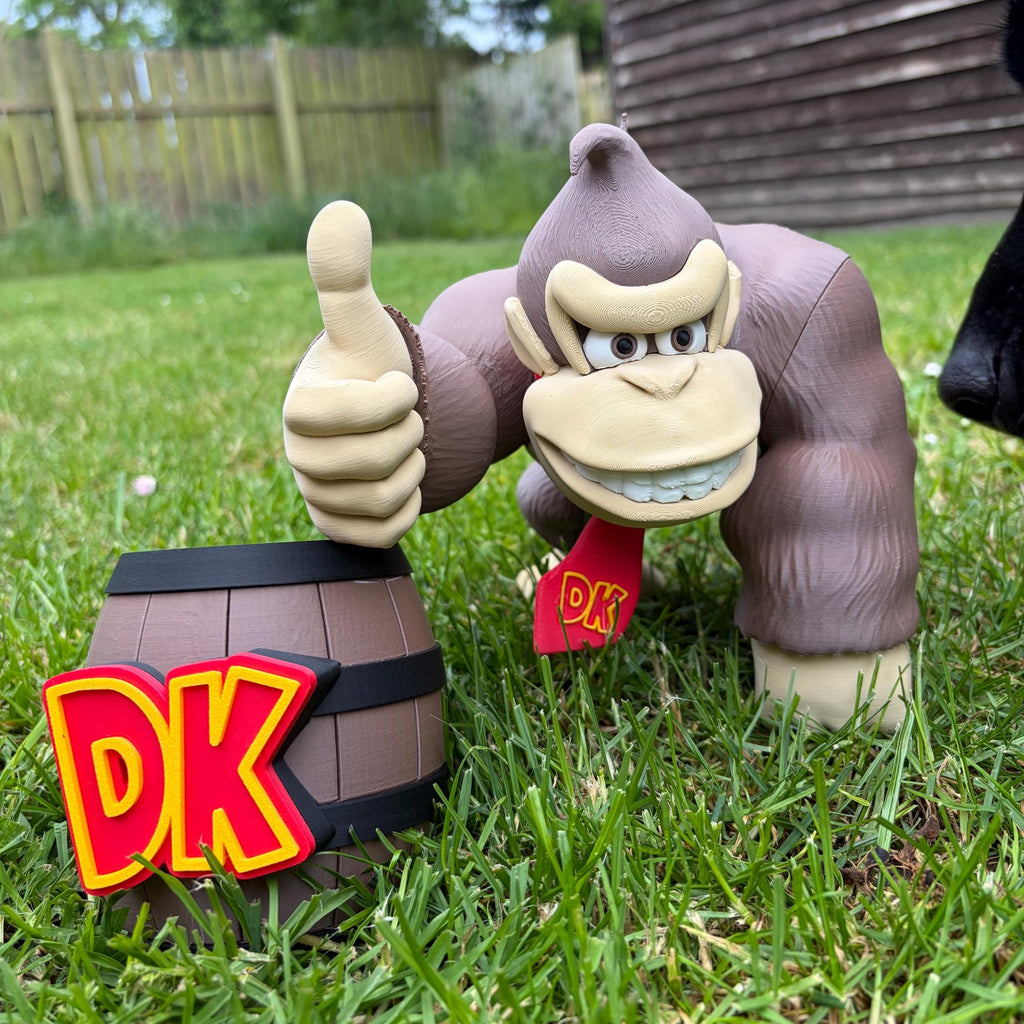 A Donkey Kong action figure posed on grass in front of a wooden shed, giving a thumbs-up beside a small barrel with a large red and yellow 'DK' sign.