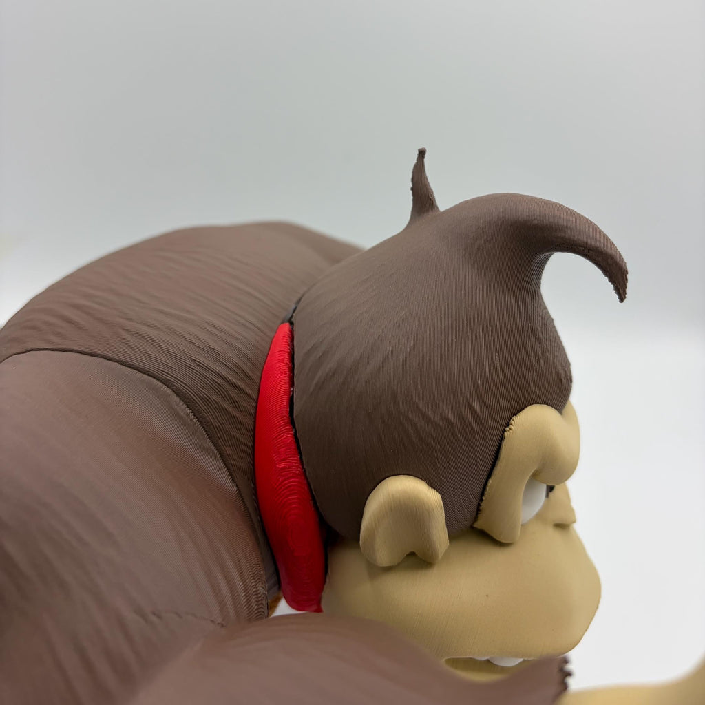 Close-up of a brown monkey figurine or toy with beige face details and a red collar, shown against a plain light gray background.