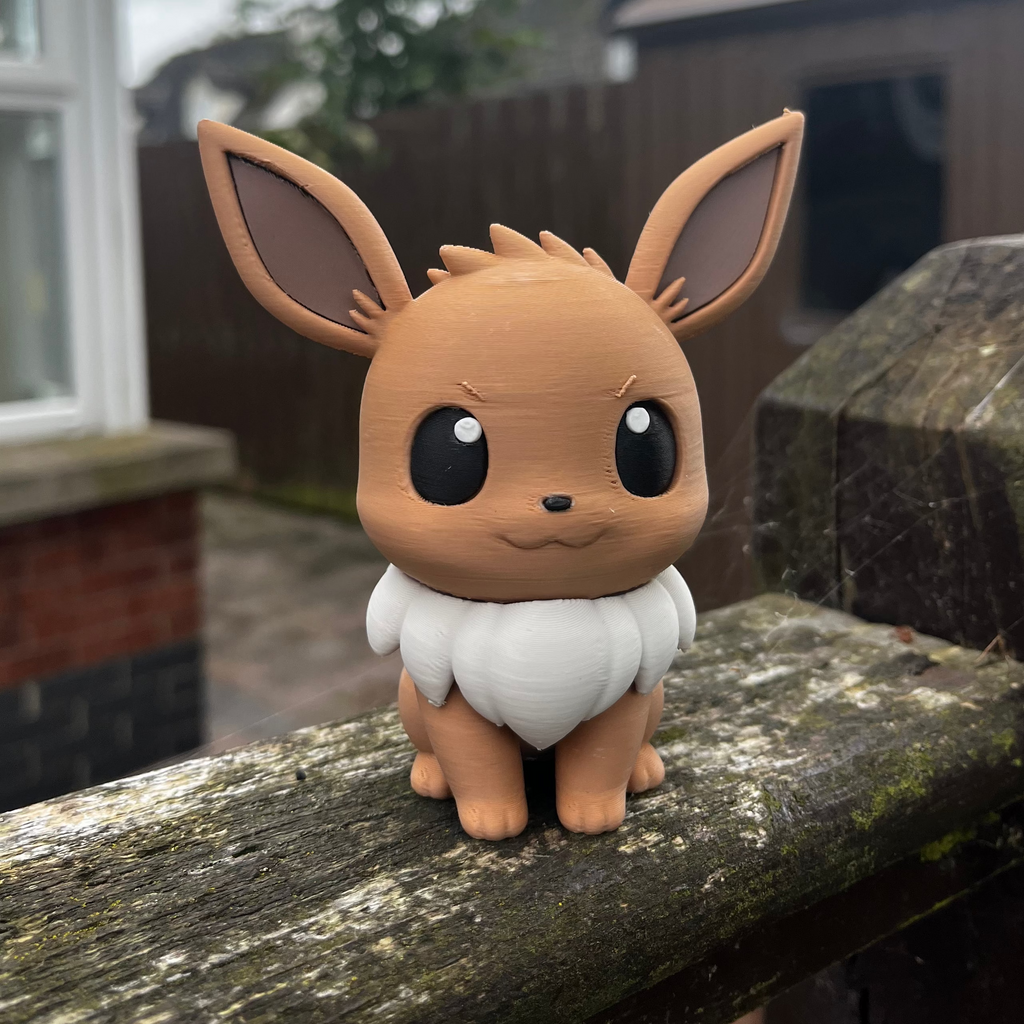 A small Eevee figurine with large black eyes, brown fur, and a white fluffy chest, sitting on a weathered wooden railing in an outdoor backyard setting.
