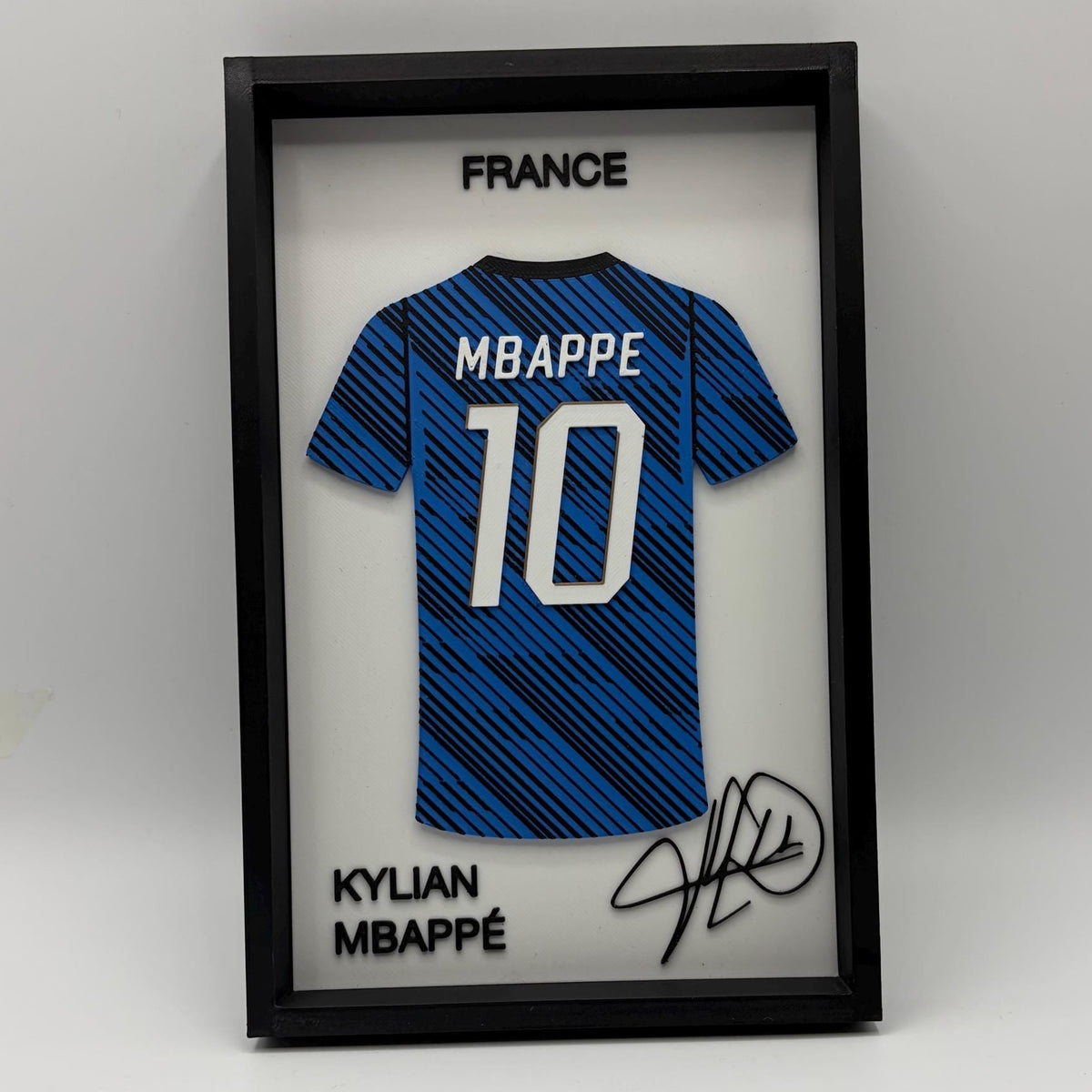 A framed Kylian Mbappé France soccer jersey display featuring a blue patterned jersey with the number 10, signed in black marker, inside a black shadowbox frame on a white background.