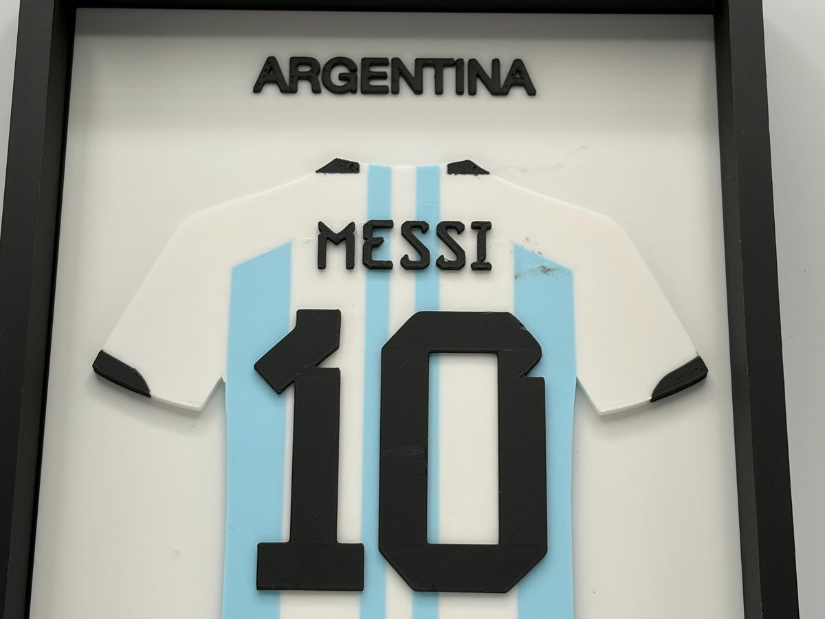 A framed 3D decorative Argentina soccer jersey featuring Lionel Messi's name and number 10. The jersey is styled with the light blue and white vertical stripes of the Argentina national team, with black text and number details.