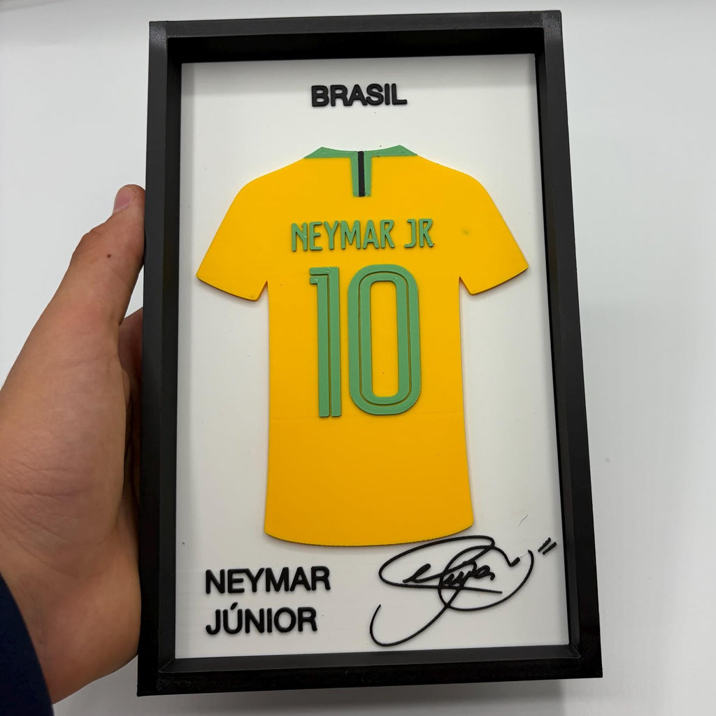 A hand holds a black-framed display featuring a yellow Brazil soccer jersey with 'NEYMAR JR' and the number '10' on the back, plus 'BRASIL' at the top and 'NEYMAR JÚNIOR' with a signature at the bottom.