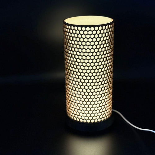 A modern cylindrical table lamp with a glowing honeycomb pattern on a dark surface against a black background.