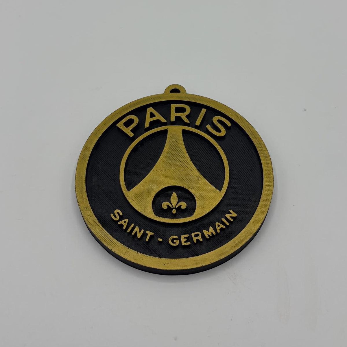 A circular Paris Saint-Germain metal pendant or charm with a gold and black design, featuring the club's logo and text, photographed on a plain light gray background.