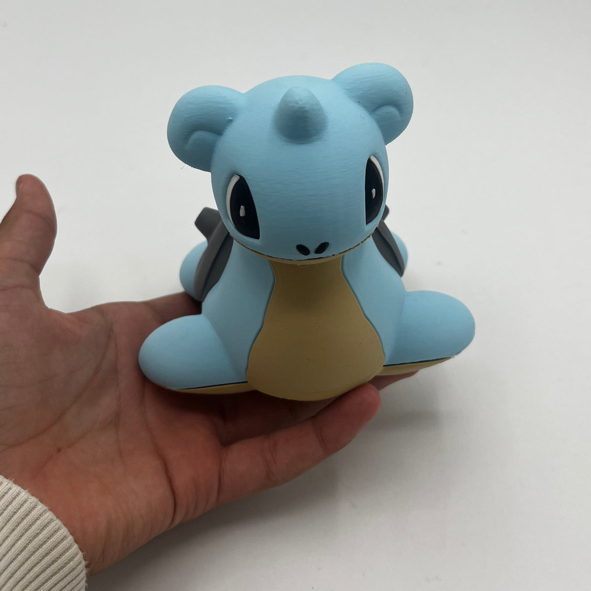 A light blue and beige toy figure being held in a person's hand against a plain white background.