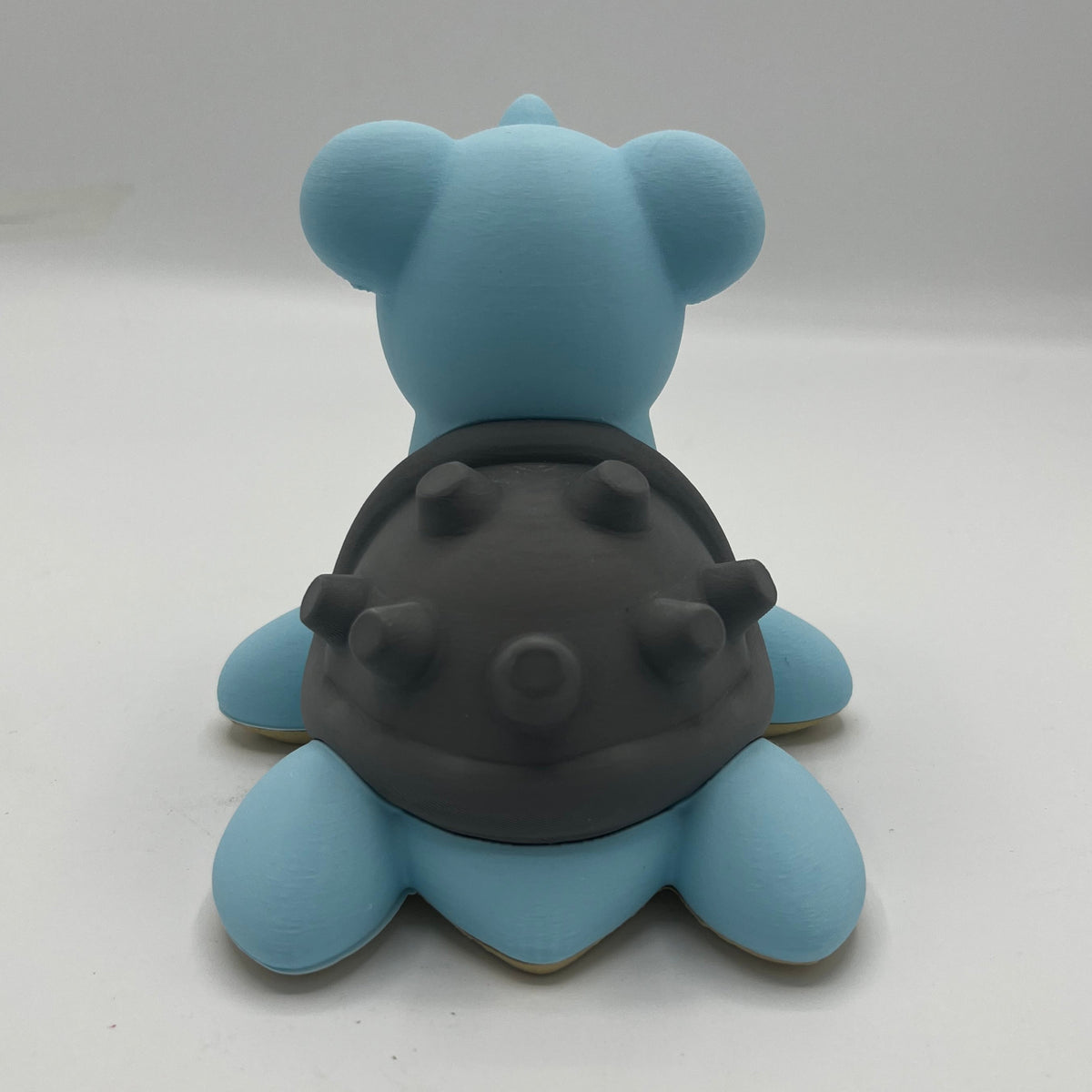 A light blue lapras figurine with a dark grey textured shell, shown from the back on a neutral light grey background.