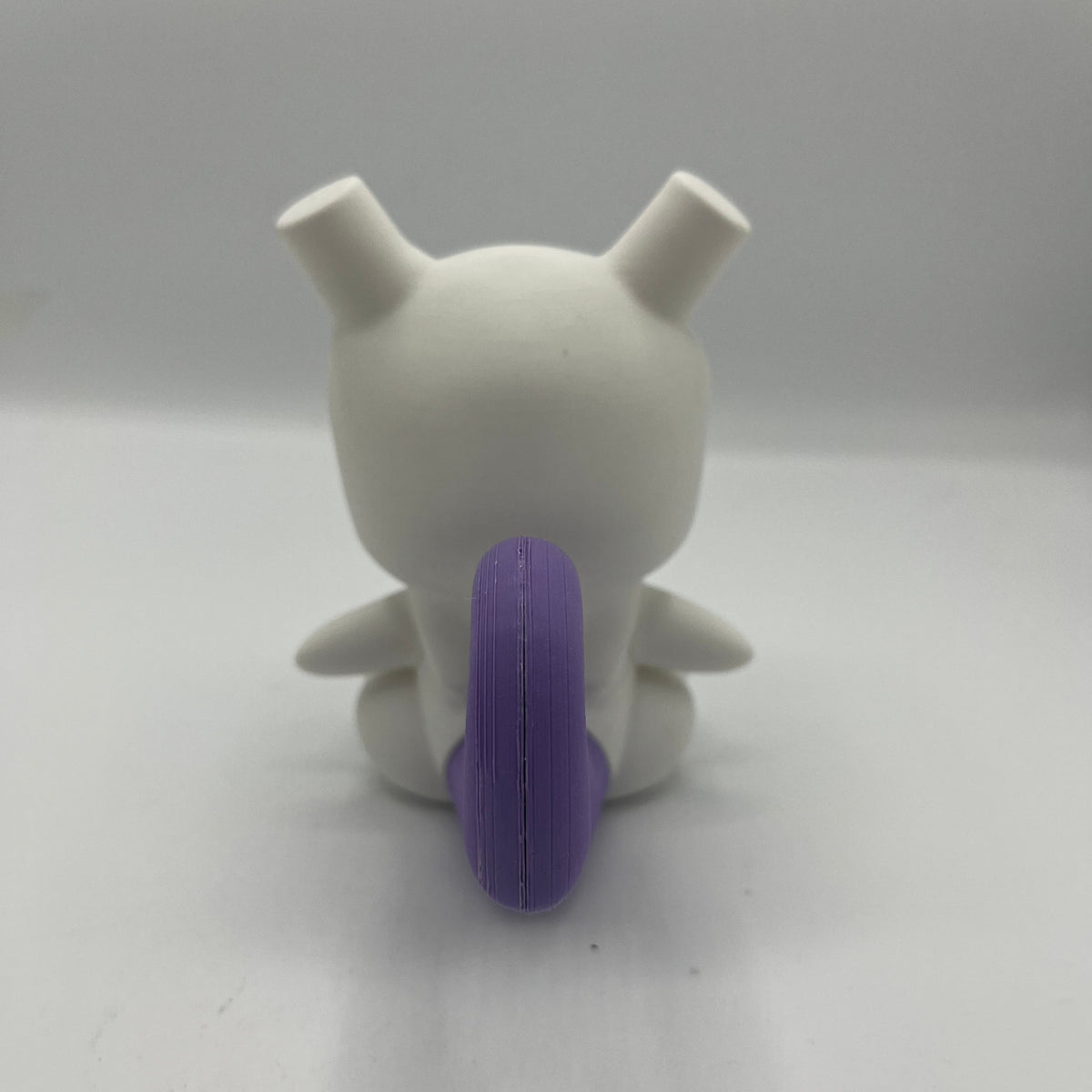 A white vinyl figure of a cartoon seen from the back, featuring a textured purple tail and small pointed ears, set against a plain background.