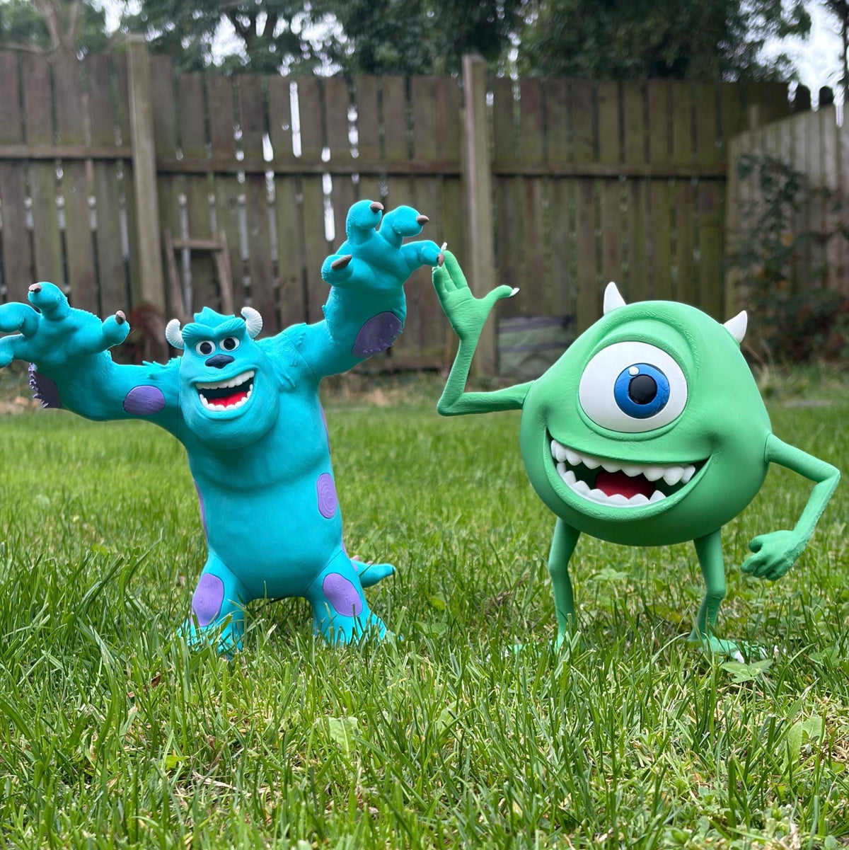 Two Mike and Sulley action figures posed outdoors on a grassy lawn in front of a wooden fence.
