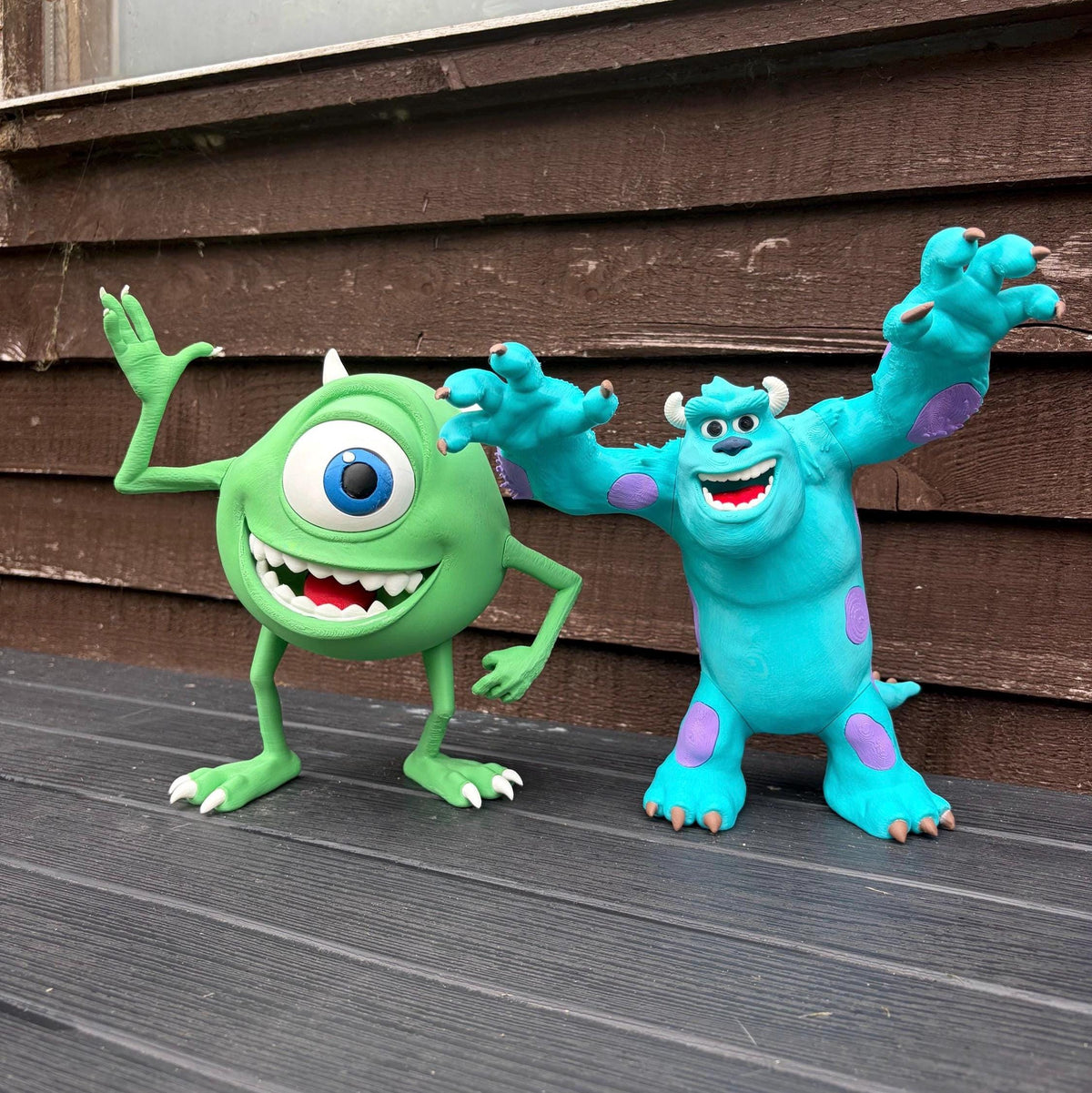 Two Mike and Sulley action figures posed together on a dark wooden deck in front of a brown wooden wall.