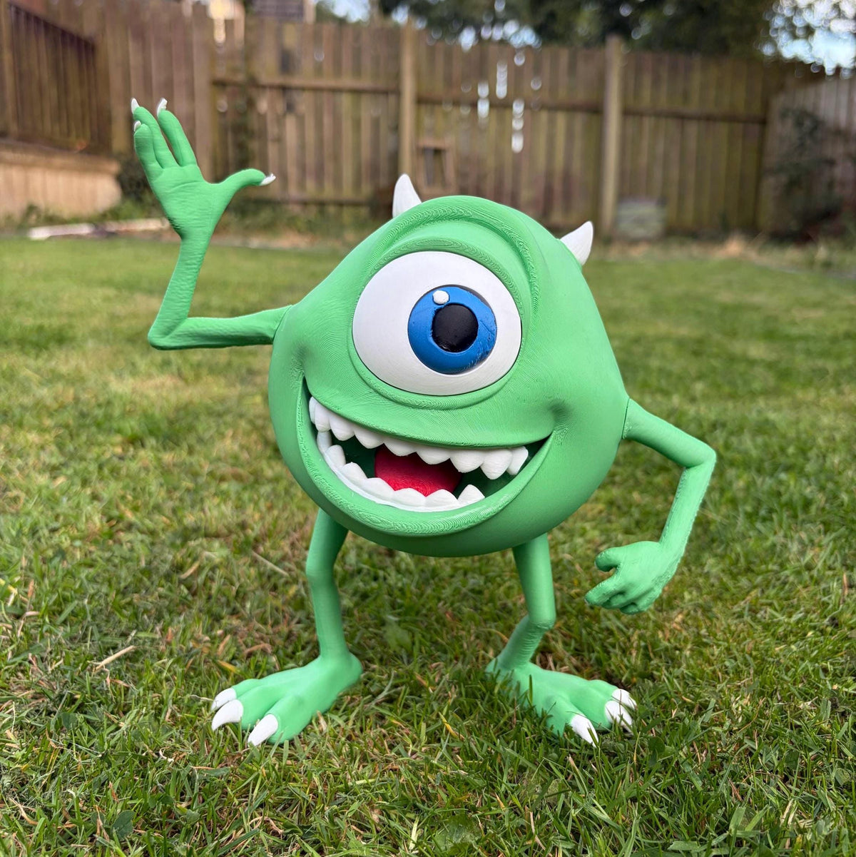 A green one-eyed cartoon character toy or figurine standing on grass in a backyard, waving with one hand and showing a wide toothy grin. The background includes a wooden fence and lawn.