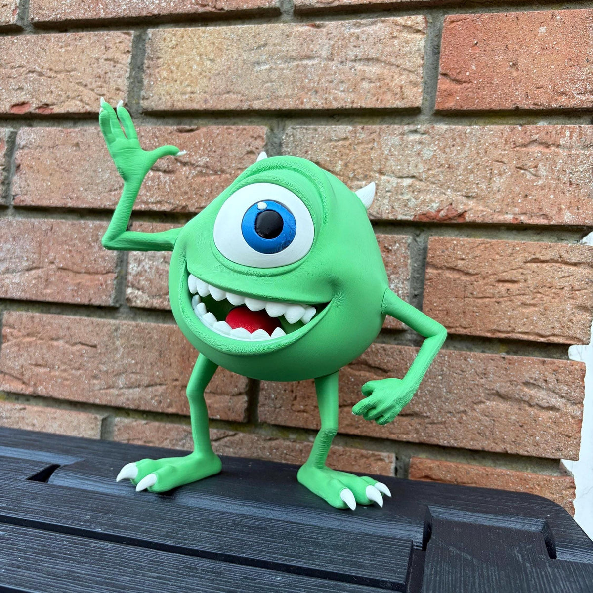 A green one-eyed Mike Wazowski figurine from Monsters Inc., standing on a dark wooden surface in front of a brick wall.