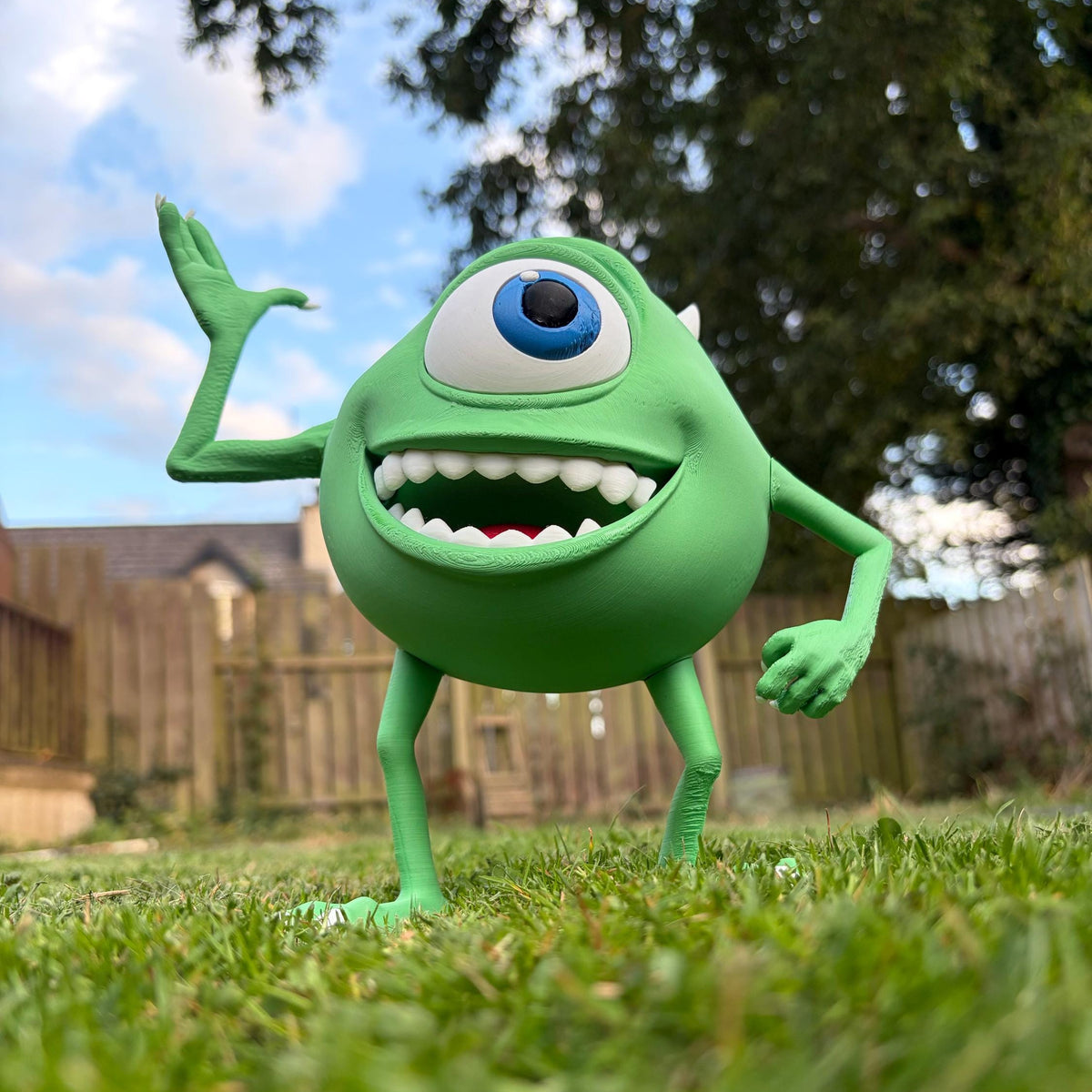 A green one-eyed cartoon character toy standing on grass outdoors, with trees, a wooden fence, and blue sky in the background.