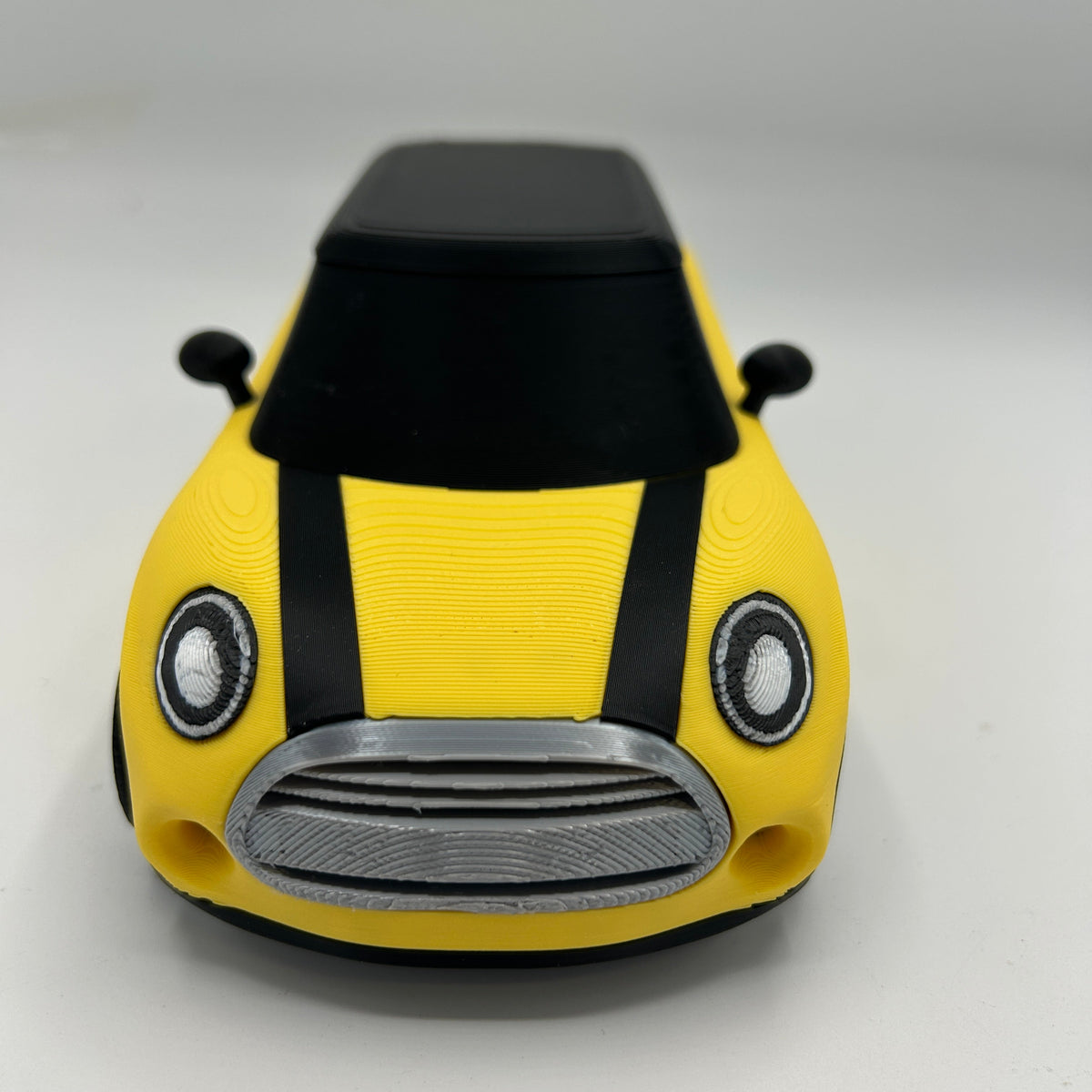 A 3D-printed toy car modeled after a Mini Cooper, featuring a bright yellow body with black roof and side mirrors, and a silver-colored front grille.