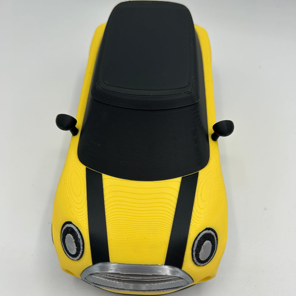 A yellow car-shaped model with a black roof, black side mirrors, and two black vertical stripes on the hood, photographed from above on a plain light background.