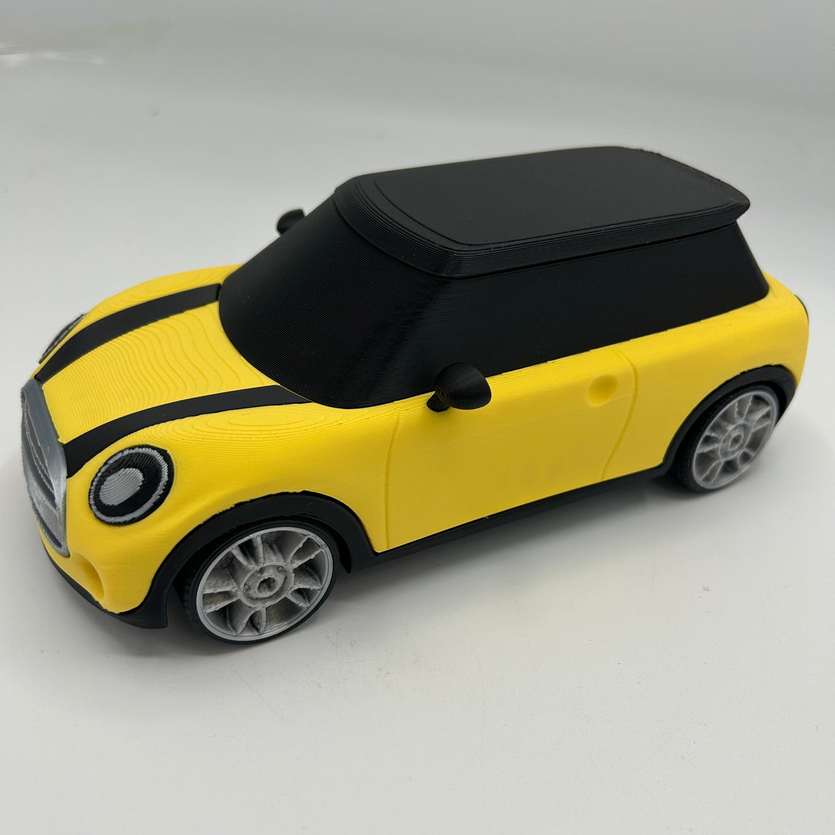 A yellow and black toy car styled like a Mini Cooper, featuring a black roof, black hood stripes, and gray wheels, photographed on a light grey background.