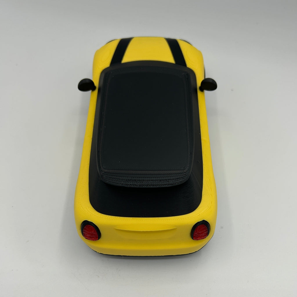A yellow toy car with a black roof and small red taillights, photographed from above on a plain light background.