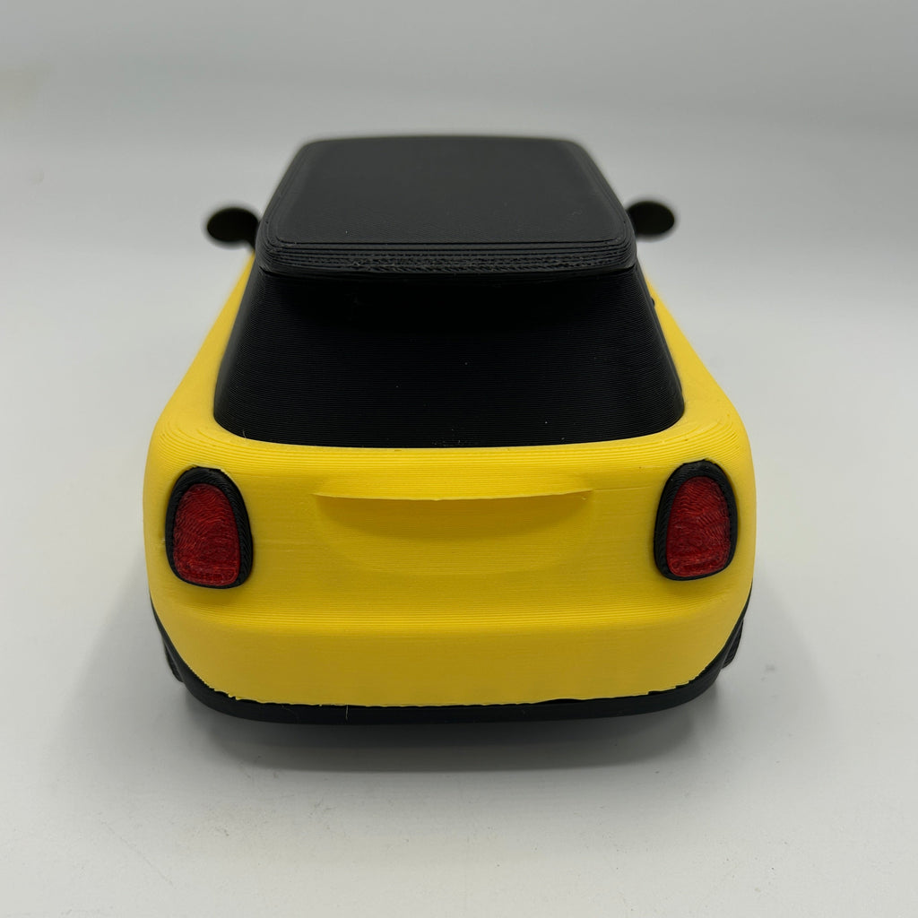 A miniature yellow toy car with a black roof and red taillights, photographed from the rear on a light gray background.