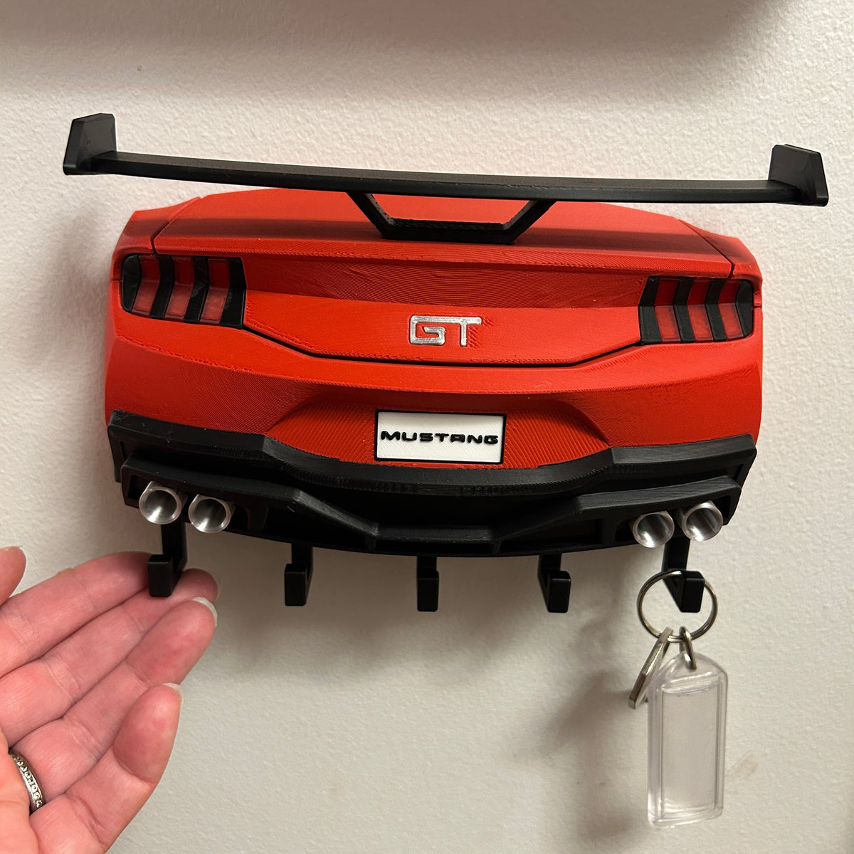 A red and black Mustang GT car trunk key holder mounted on a beige wall, with a hand holding one of the black hooks on the left and keys hanging from the right.