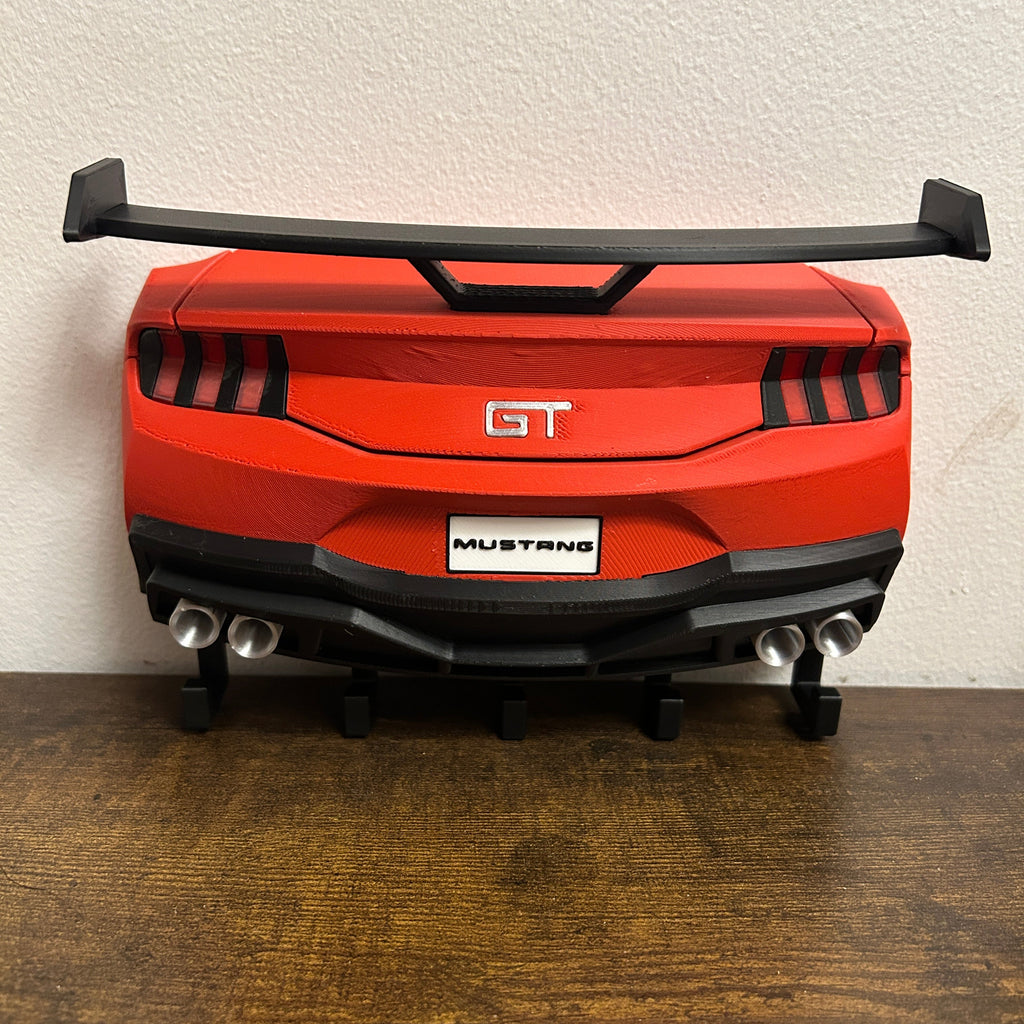 A red and black toy model of a Ford Mustang GT car rear, displayed on a wooden surface against a textured off-white wall.