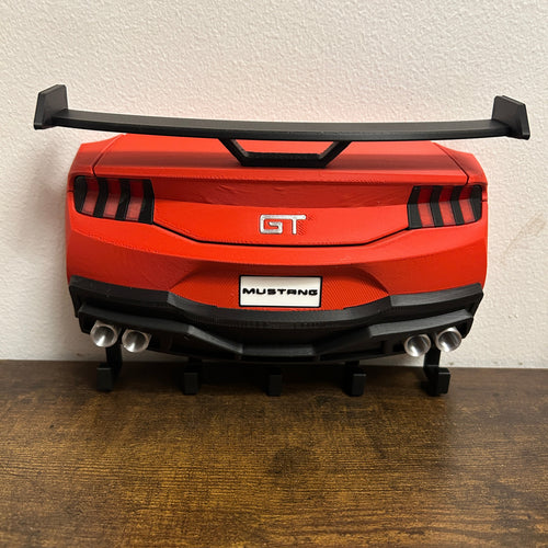 A red and black toy model of a Ford Mustang GT car rear, displayed on a wooden surface against a textured off-white wall.