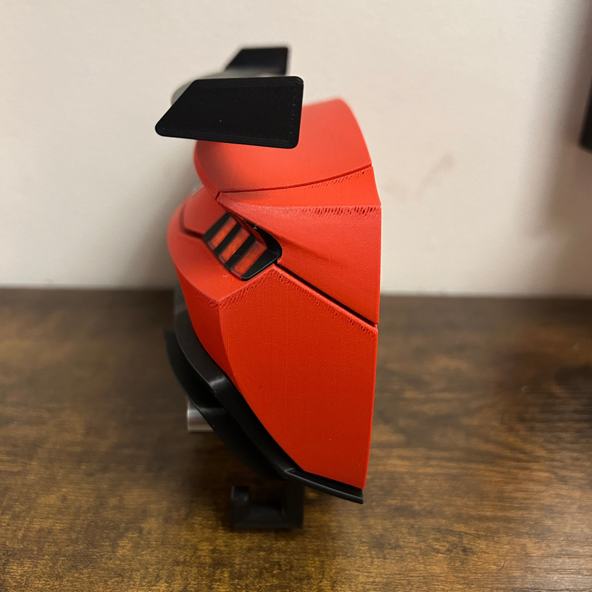 A red and black car key holder accessory displayed on a small stand, set on a wooden tabletop against a plain off-white wall.