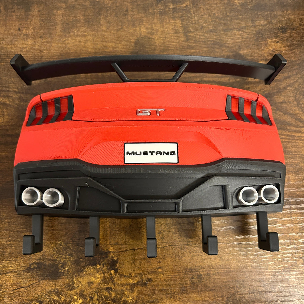 A red and black Mustang rear spoiler wing accessory with a white Mustang badge, displayed on a wooden table.