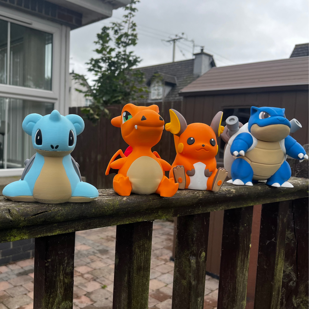 Four cute toy figurines resembling popular cartoon Pokémon characters are lined up on a weathered wooden railing in front of a residential house. The toys feature a variety of colours including brown, light blue, orange, and white.