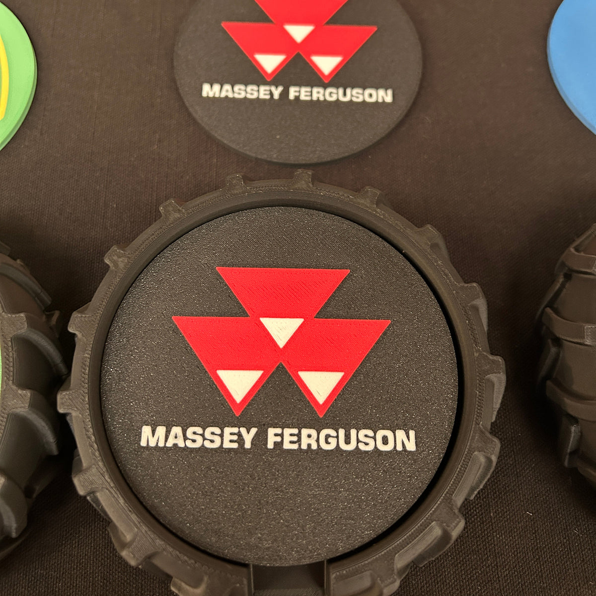 A close-up of a black textured foam disc with a red and white Massey Ferguson logo and white brand text, shown on a dark surface with other colorful discs partially visible around it.