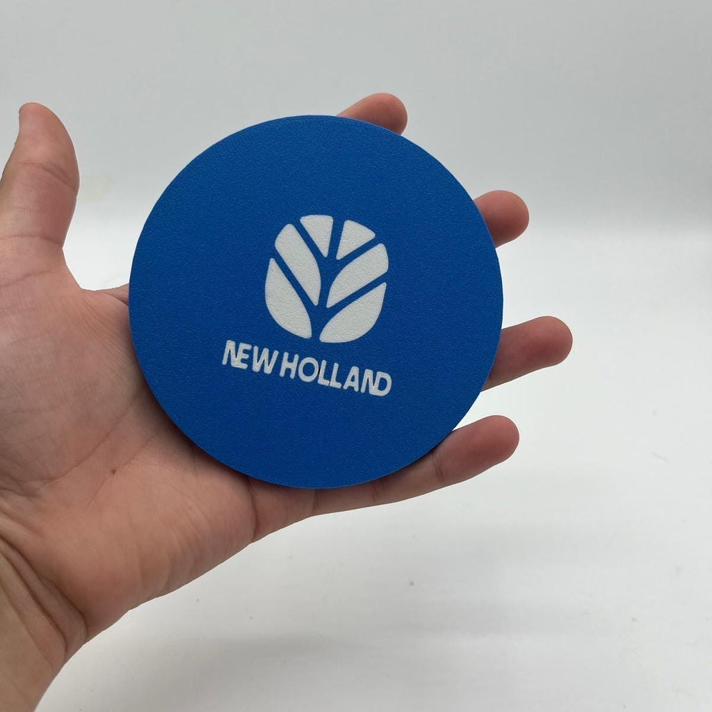 A hand holding a round blue coaster with a white leaf logo and the text 'NEW HOLLAND' printed on it, set against a plain light gray background.