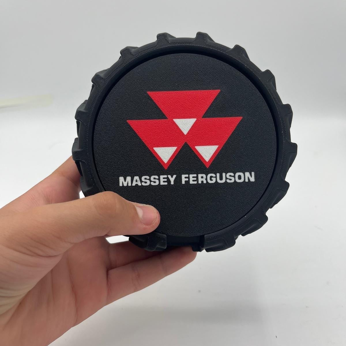 A hand holding a black circular tyre with a red and white Massey Ferguson logo coaster and white text, against a plain light grey background.