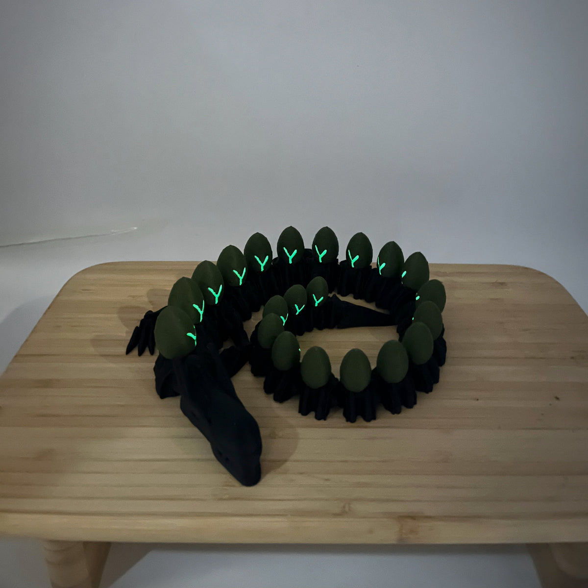 A coiled black and olive green snake-like accessory with glowing green accents, displayed on a light wood table against a plain light grey background.