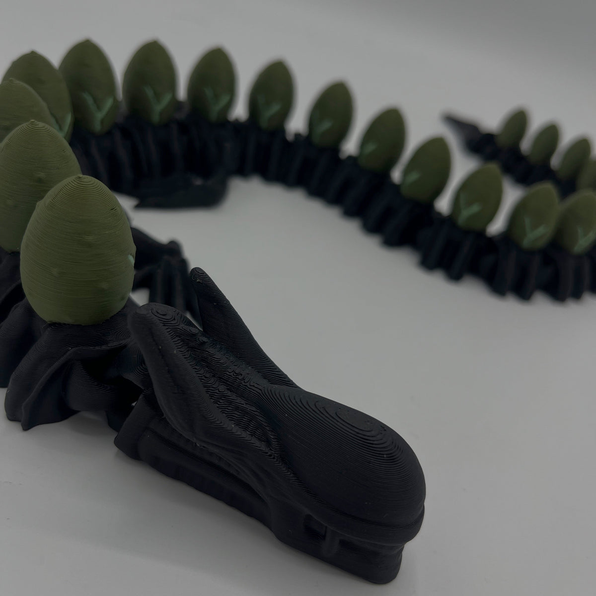 A black 3D-printed snake head toy with olive green scales along its back, shown on a plain light grey background.