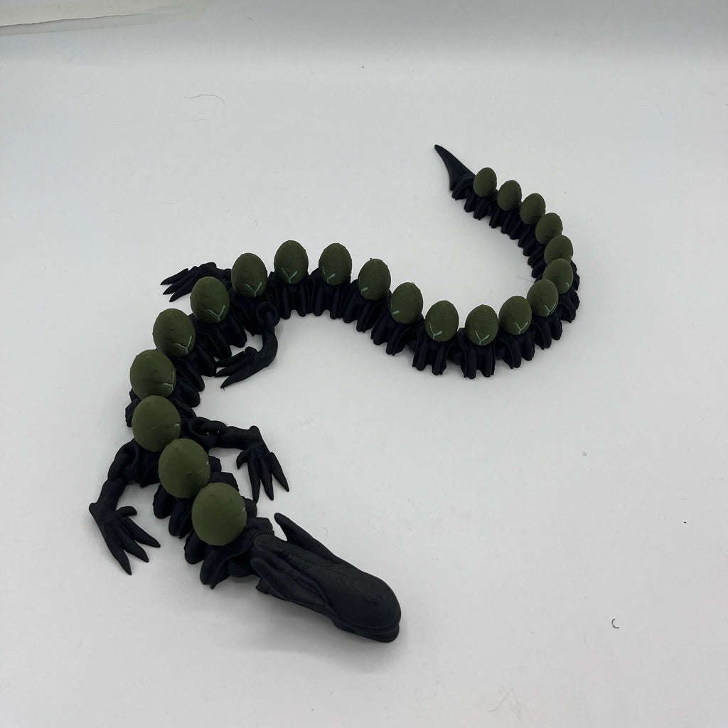 A 3D-printed articulated toy alligator or crocodile, shown coiled on a plain white background. The toy features a black body with olive green circular plates along its back and head.