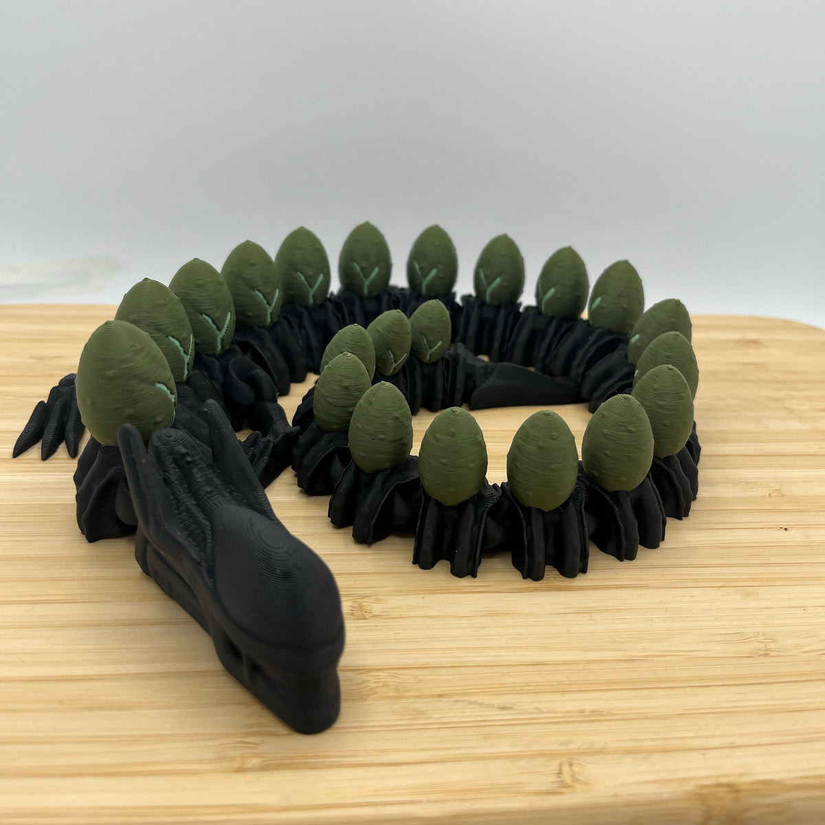 A black dragon figurine with green scales, displayed on a light wood cutting board against a plain white background.